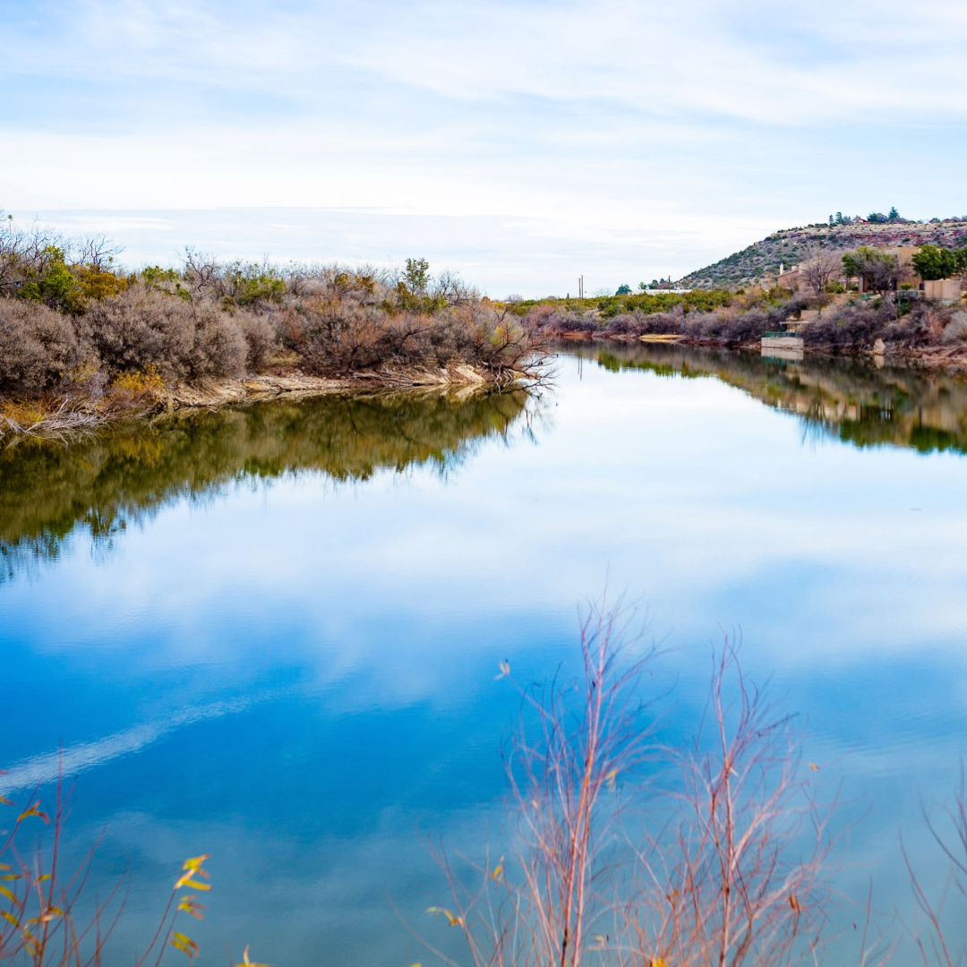 Calm blue waters of Comanche Trail Lake mirror the clear sky above, bordered by shrubs and trees.